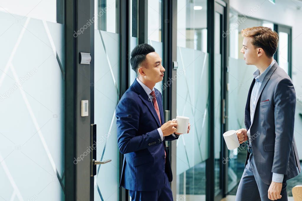 Serious young stylish Asian businessman with mig of coffee looking at his colleague telling news and his creative ideas