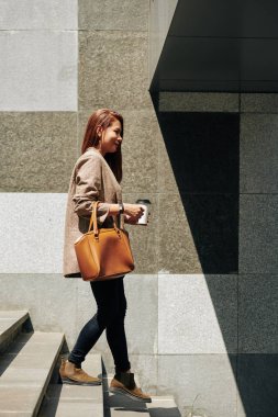 Kahve fincanı taşıyan neşeli genç bir kadın metroya doğru yürüyor.