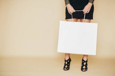 Yüksek topuklu bir kadının, elinde pahalı bir şeyle büyük beyaz bir alışveriş çantasıyla dikilirken görüntüsü.