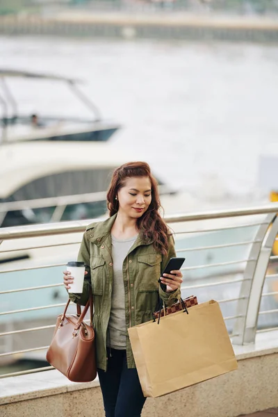微笑着带着购物袋散步的女性旅行者 并在智能手机上查看社交媒体 — 图库照片