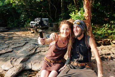 Ormanda taşın üzerinde oturan ve Monopod 'a bağlı akıllı telefondan selfie çeken mutlu olgun bir çift.