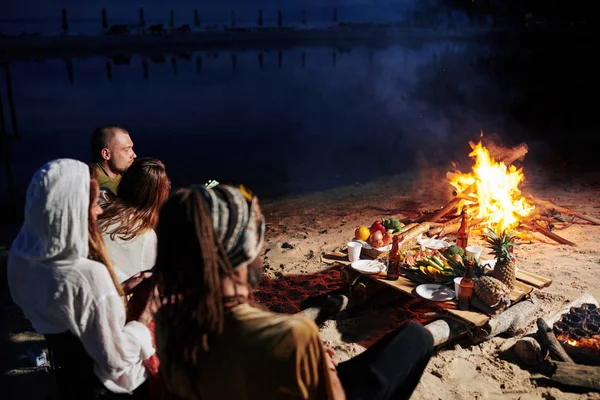 Friends resting on beach in the evening, sitting by fire and eating fresh fruits and seafood