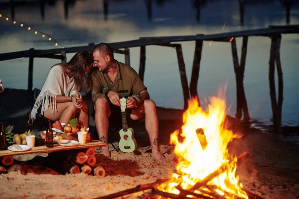 Śmiejąca Się Młoda Para Siedząca Przy Ogniu Plaży Grająca Ukulele — Zdjęcie stockowe