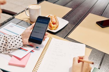 İş kadını kafe masasında oturuyor ve iş seyahatini planlarken akıllı telefon uygulaması ile banka hesabını kontrol ediyor.