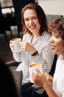 Polka noktalı bluzlu çekici genç kadın kahve içiyor ve arkadaşının komik hikayesini dinlerken gülüyor.