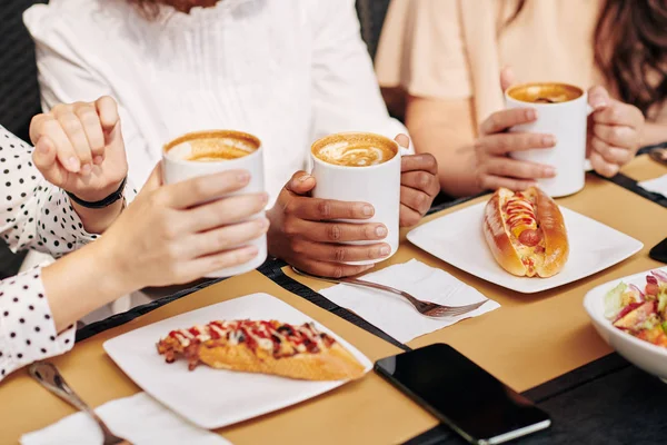 Female friends or colleagues drinking coffee and eating hotdogs at lunch in cafe