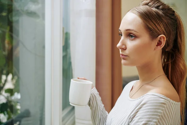 深刻な若いですブロンド女性飲むコーヒーのマグカップと窓を通して見る — ストック写真
