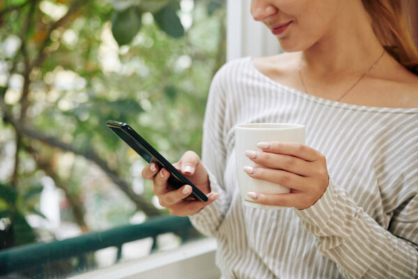 Cropped image of smiling young woman drinking cup of coffee and checking text messages in smartphone
