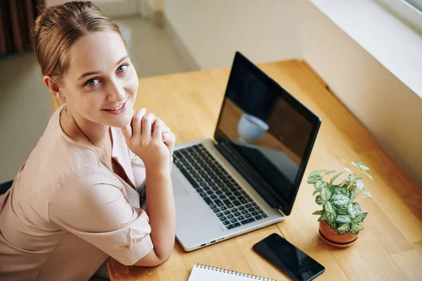 Horizontal high angle shot of attractive Caucasian woman sitting at table with laptop on it looking at camera smiling