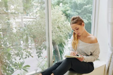 Romantik, beyaz bir kızın pencere eşiğinde oturup defterine bir şeyler yazarken çekilmiş yatay bir fotoğrafı.