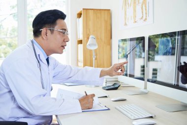 Masada oturmuş bilgisayardaki röntgen görüntülerini inceleyen erkek doktorun yatay yan görüntüsü.