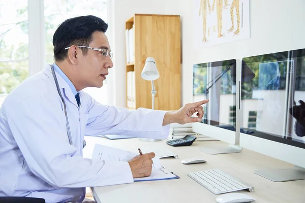 コンピュータ上のX線画像を調べる机に座っている男性医師の横側のビューショット — ストック写真