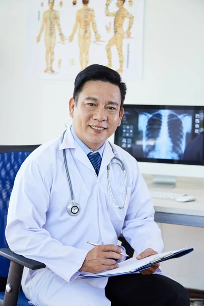 Mature Asian Doctor Sitting Chair Holding Medical Record Looking Camera — Φωτογραφία Αρχείου
