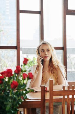 Modern kafede tek başına oturmuş telefonla konuşan şık bir genç kadının dikey çekimi.
