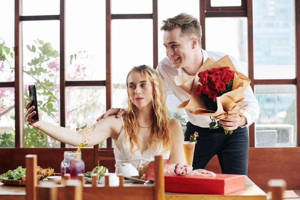 在现代咖啡馆里 一个快乐的年轻男子和一个迷人的年轻女子带着一束红玫瑰在约会时的横向照片 — 图库照片
