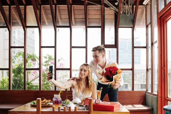 在现代咖啡馆里 年轻英俊的男人和迷人的女人拿着一束红玫瑰自拍的水平镜头 — 图库照片