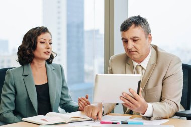 Orta yaşlı beyaz bir adam ve olgun bir kadın ofis masasında birlikte çalışıyor, yatay çekimde.