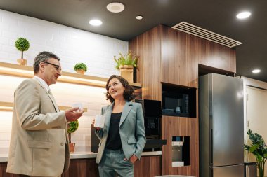 Kendine güveni tam bir erkek ve şık takım elbiseli güzel bir kadın kahve molası sırasında sohbet ediyorlar. Mutfakta duruyorlar.