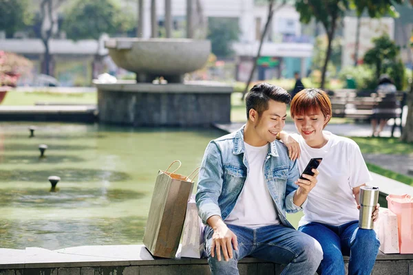 纵横拍年轻男女和购物袋坐在一起 面对着喷泉 用智能手机观看某件事情 — 图库照片