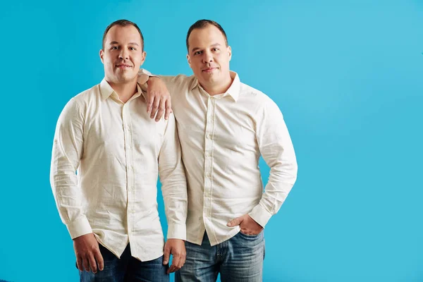 两个穿着白衬衫和牛仔裤的同卵双胞胎站在蓝色背景下看着摄像机 — 图库照片