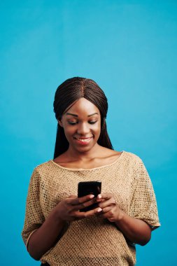 Stüdyo portresi, akıllı telefonuyla internette sörf yapan çekici siyahi kadın gülümsemesi, mavi arka plan.