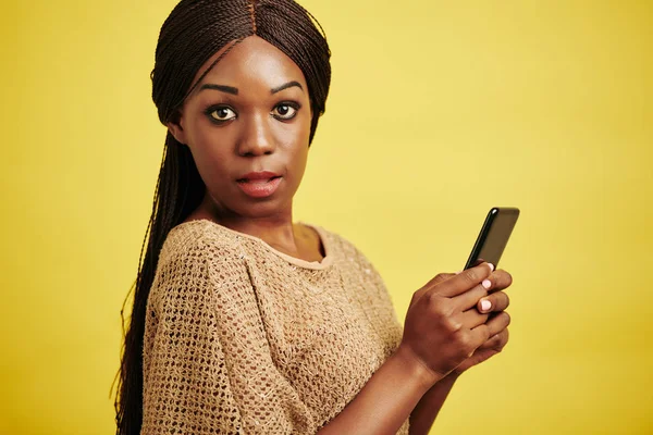 カメラを見てスマートフォンを保持している魅力的な黒の女性の水平スタジオの肖像画 黄色の背景 — ストック写真