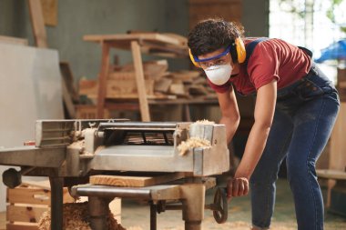 Ahşap işleme. Maskeli, güvenlik gözlüklü ve kulaklıklı kadın marangoz ahşap işleme makinesinde ahşap tahta işliyor.