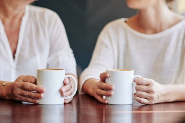 Yetişkin bir kadın ve yetişkin kızının kahve içip evde haberleri ve dedikoduları tartışırken görüntüsü.