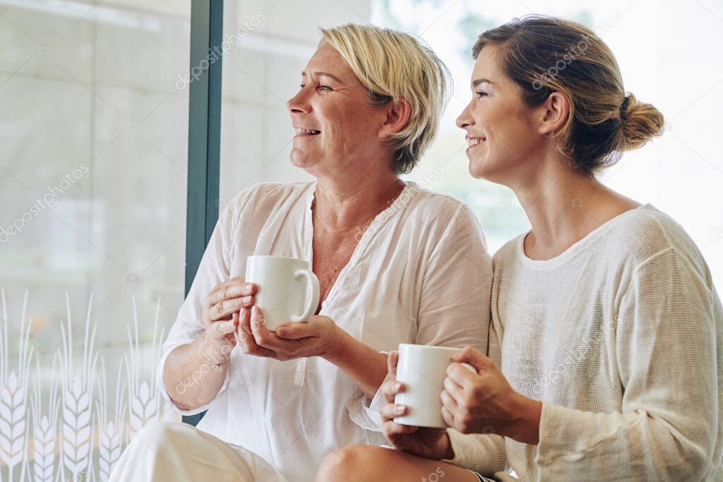 Happy middle-aged woman and her adult daughter drinking morning coffee and looking through big window