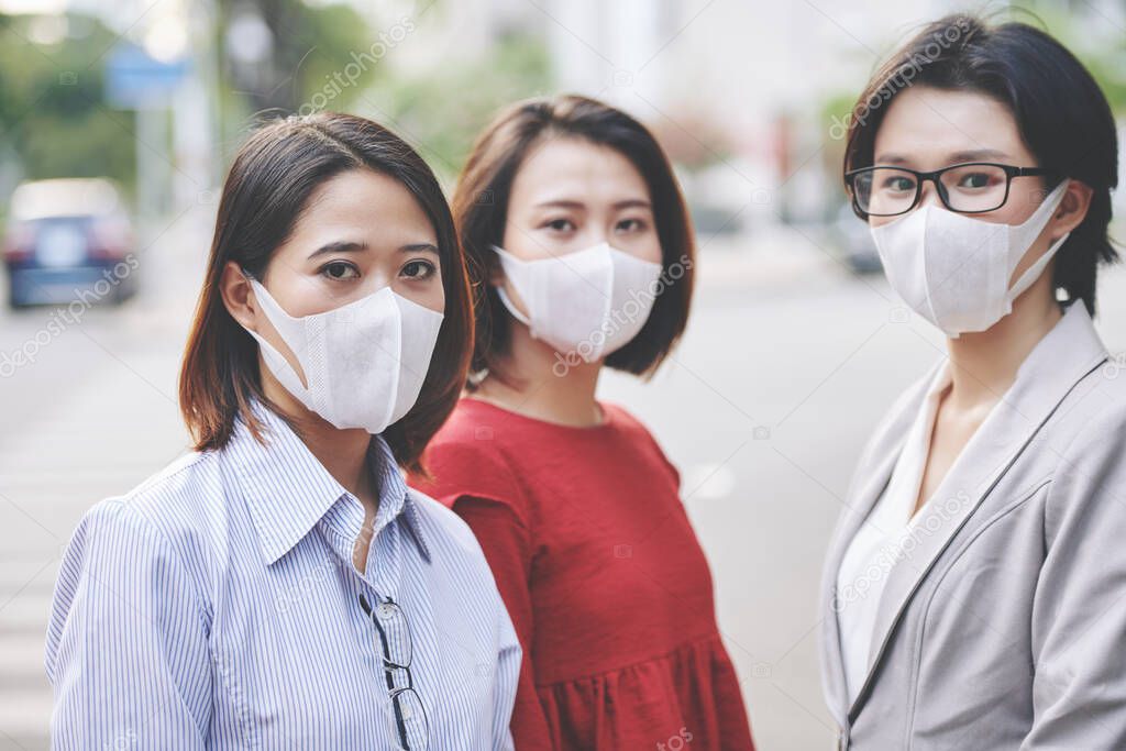 Group of serious young Asian women in antibacterial masks stranding in city street