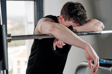Spor salonunda halterlere yaslanmış yorgun ve formda genç bir adam.