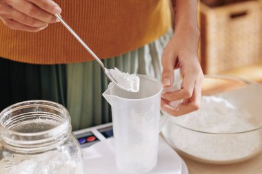 Evde sabun yaparken plastik kavanoza kaşık sodyum sodyum sodyum sodyum sodyumu koyan kadının yakın plan resmi.