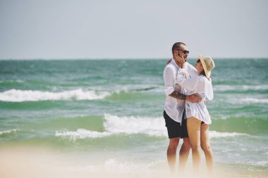 Mutlu genç çift plajda ıslak beyaz gömleklerle durup birbirlerine bakıyorlar.