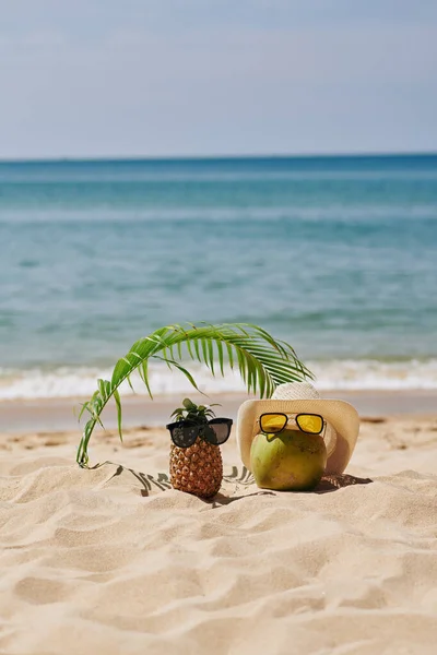 ビーチのヤシの葉の下で休んでいるサングラスのココナッツとパイナップル — ストック写真