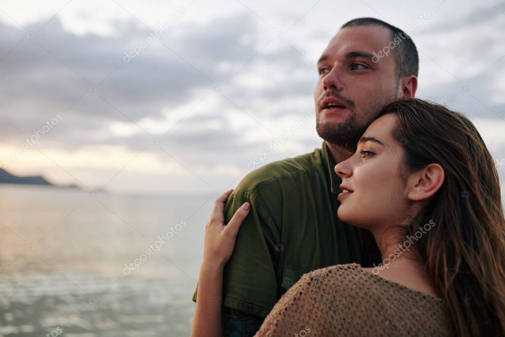 Hugging young couple standing on beach and looking at beautiful seascape