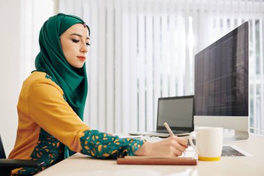 Müslüman genç kadın yeni proje için programlama kodu üzerinde çalışırken planlamacıda notlar alıyor