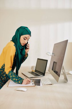 Musilim yazılım mühendisi proje yöneticisi ile telefonda konuşuyor ve fikirleri not defterine yazıyor.