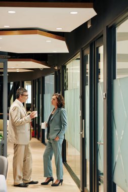 Olgun iş adamları kahve kupaları ile ofis koridorunda durup önemli konuları tartışıyor.