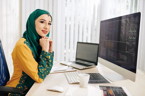 Beautiful young muslim woman in hijab sitting at office table in front on computer with programming code on the screen