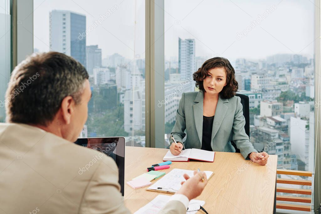 Beautiful confident business lady looking at finance department head talking about estimated sales and taking notes in planner