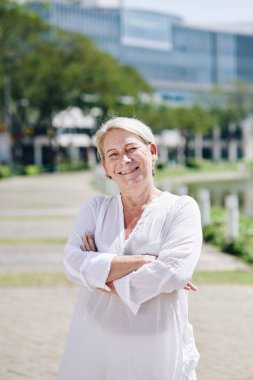 Kollarını kavuşturup kameraya bakan, gülümseyen olgun bir kadının portresi.