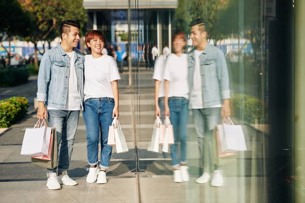 若いアジアのカップルが紙袋を持って通りを歩き お店の窓を見て笑っています — ストック写真