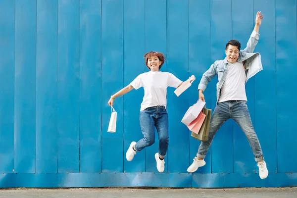 Opgewonden Jong Stel Met Boodschappentassen Springt Tegen Blauwe Muur — Stockfoto