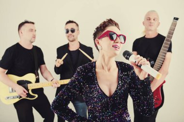 Müzik grubu arka planda çalarken, güneş gözlüklü genç bayan şarkıcı mikrofonla şarkı söylüyor.