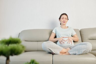 Güzel gülümseyen genç Vietnamlı kadın evdeki kanepede meditasyon yaparken karnına dokunuyor.