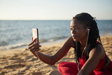 Koşu yaptıktan ve aboneleri için video çektikten sonra plajda dinlenen pozitif, güzel bir genç bayan.