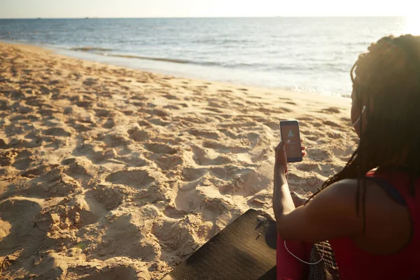 砂浜に座っている若い女性を瞑想し スマートフォンアプリ経由でガイド付きセッションに耳を傾ける — ストック写真