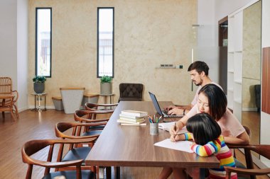 Genç adam dizüstü bilgisayarda çalışırken karısı kızıyla bir şeyler çizip ona yazmayı öğretiyor.