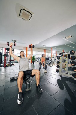 Spor salonunda ağırlık çalışan insanlar, erkek dambılları kaldırırken çömelen genç bir kadın.
