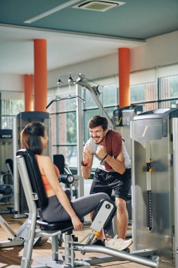 Heyecanlı duygusal fitness eğitmeni iç ve dış bacak makinesinde egzersiz yapan genç kadını motive ediyor ve cesaretlendiriyor.